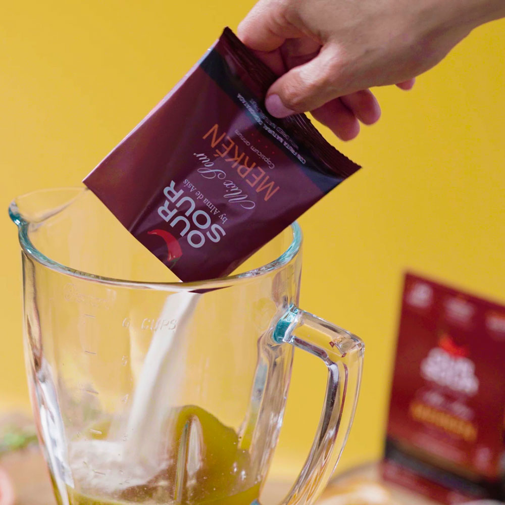 Preparando cóctel sour con polvo de limón y merkén Our Sour. Mano vertiendo un sachet en una licuadora, con fondo amarillo y empaque desenfocado al fondo